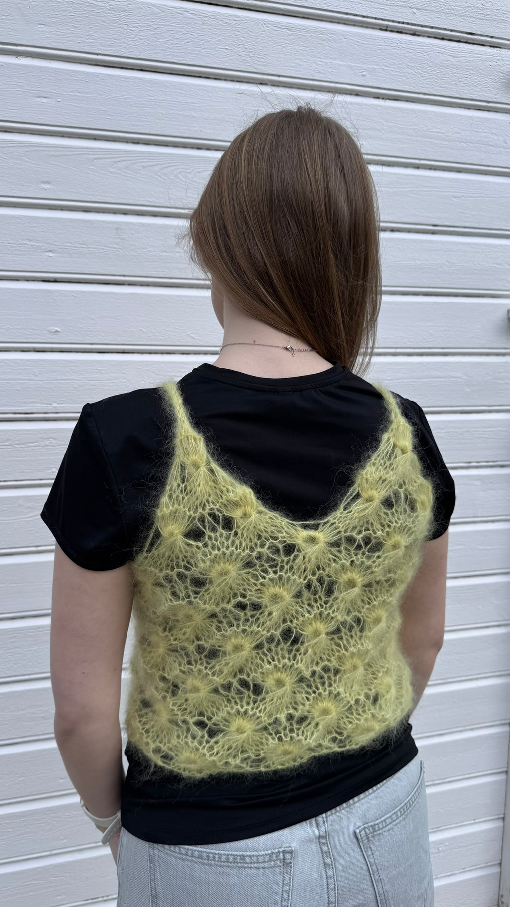 Person wearing a black t-shirt with a knitted yellow lace vest over it against a white wall.