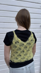 Person wearing a black t-shirt with a knitted yellow lace vest over it against a white wall.