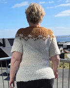 Woman standing on a balcony overlooking a coastal view with mountains in the distance. She's wearing a white and brown knitted sweater.