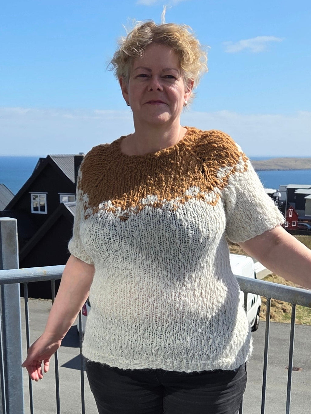 Woman standing on a metal staircase with a clear blue sky and some greenery in the background. She's wearing a white and brown knitted blouse.