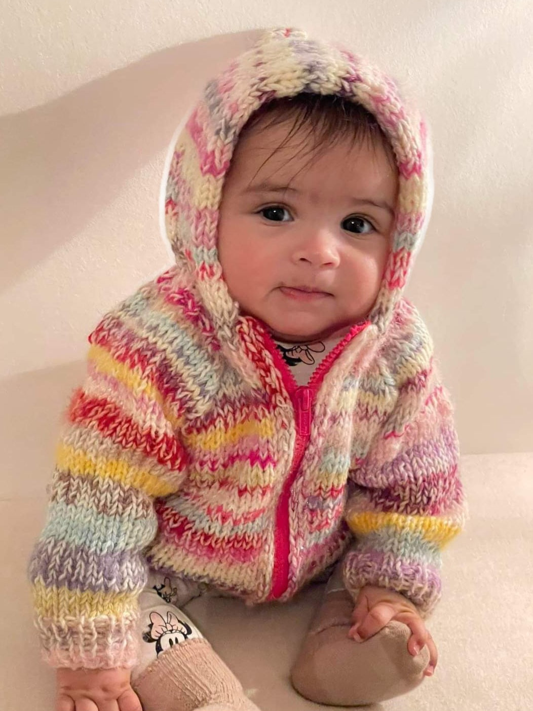 Baby wearing a colorful knitted hoodie sitting on a white surface.