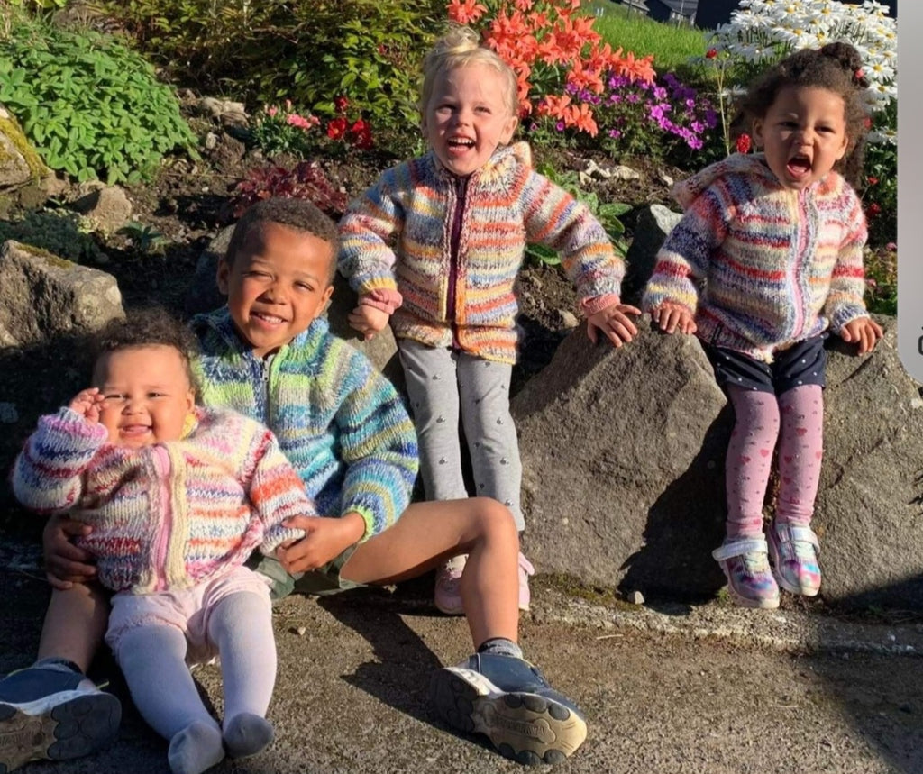 Four children in colorful, knitted hoodies sitting on rocks with flowers in the background