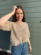Woman wearing a beige sweater with colorful sequins and blue jeans against a gray wooden background
