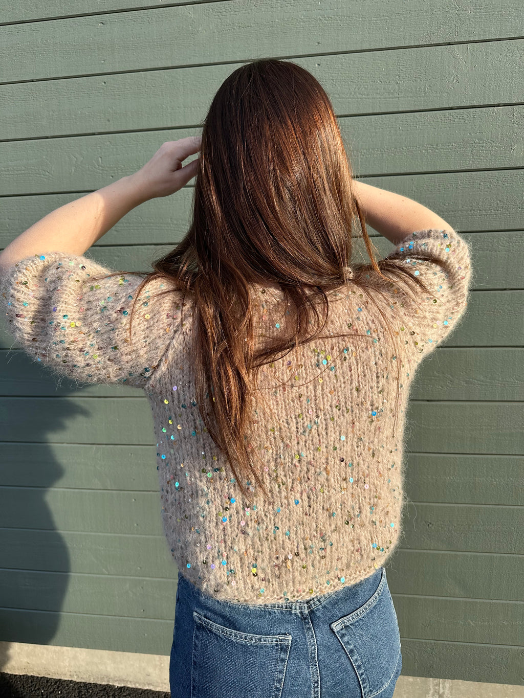 Woman wearing a sequined sweater and blue jeans against a green wooden background