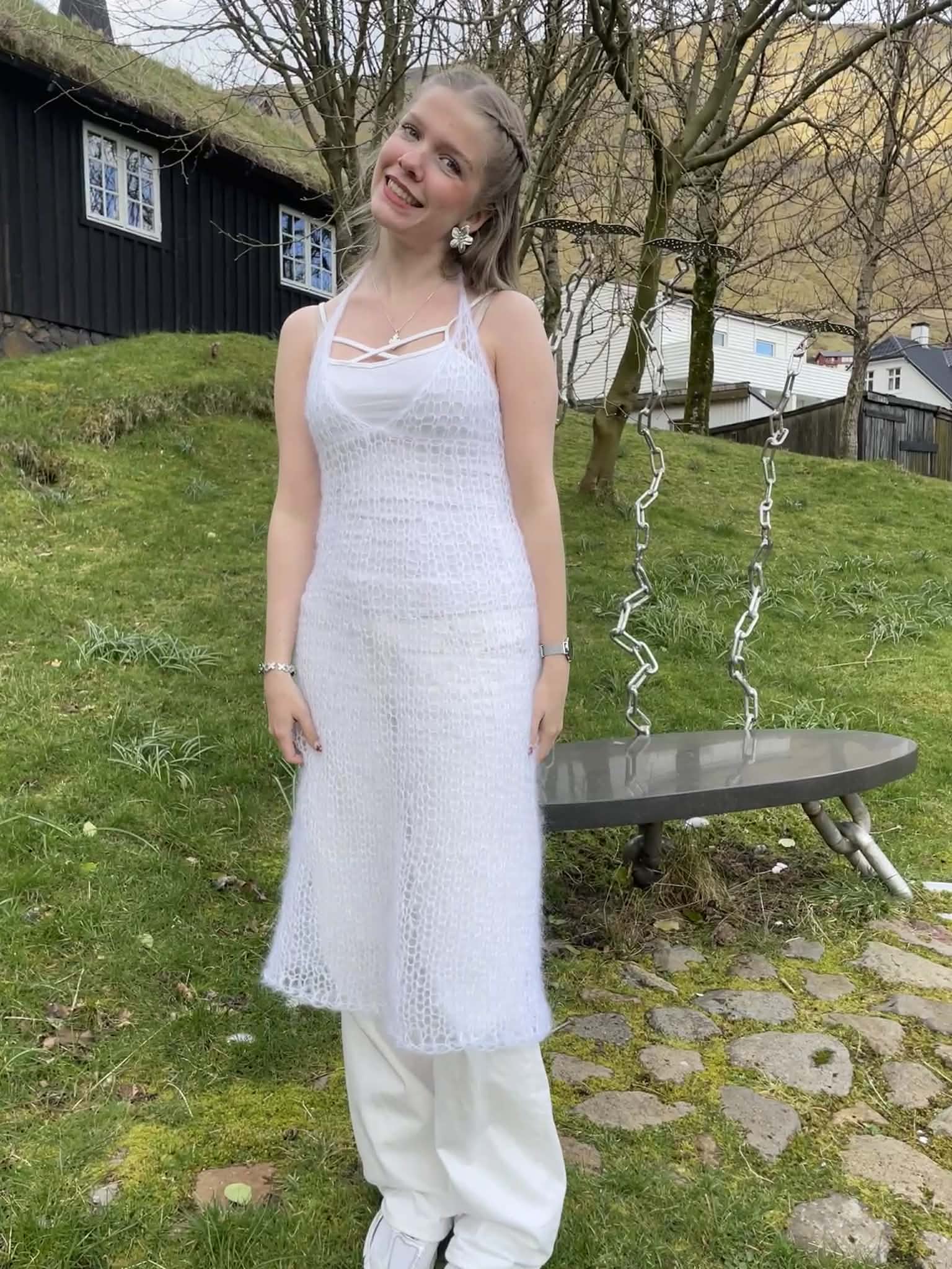 Woman in a white, knitted dress standing outdoors with a bench and trees in the background