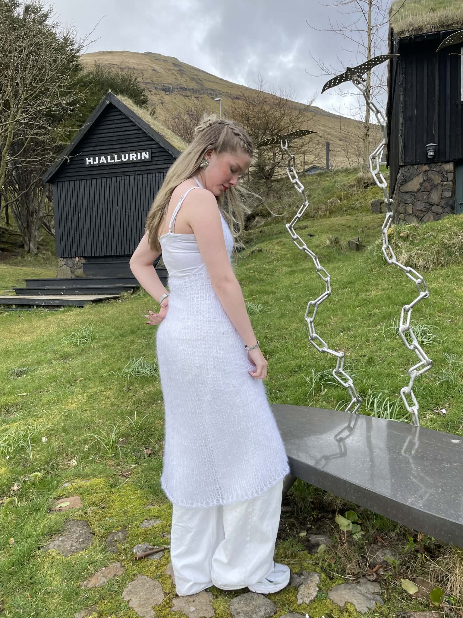 Woman in a white, knitted dress standing in a grassy area with a black building and mountains in the background.