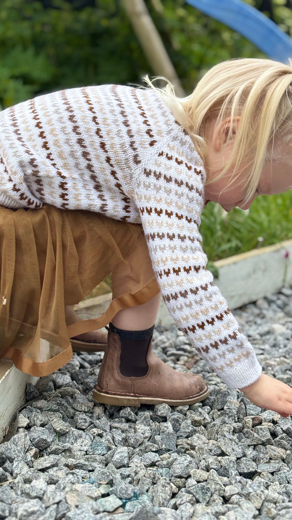 Child wearing a patterned sweater and skirt on a gravel surface with greenery in the background
