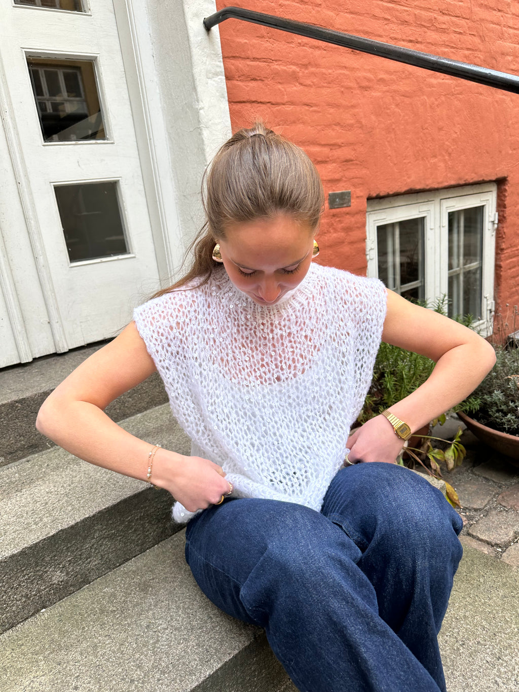 Woman sitting on steps wearing a white lace top and blue jeans, with a building in the background.