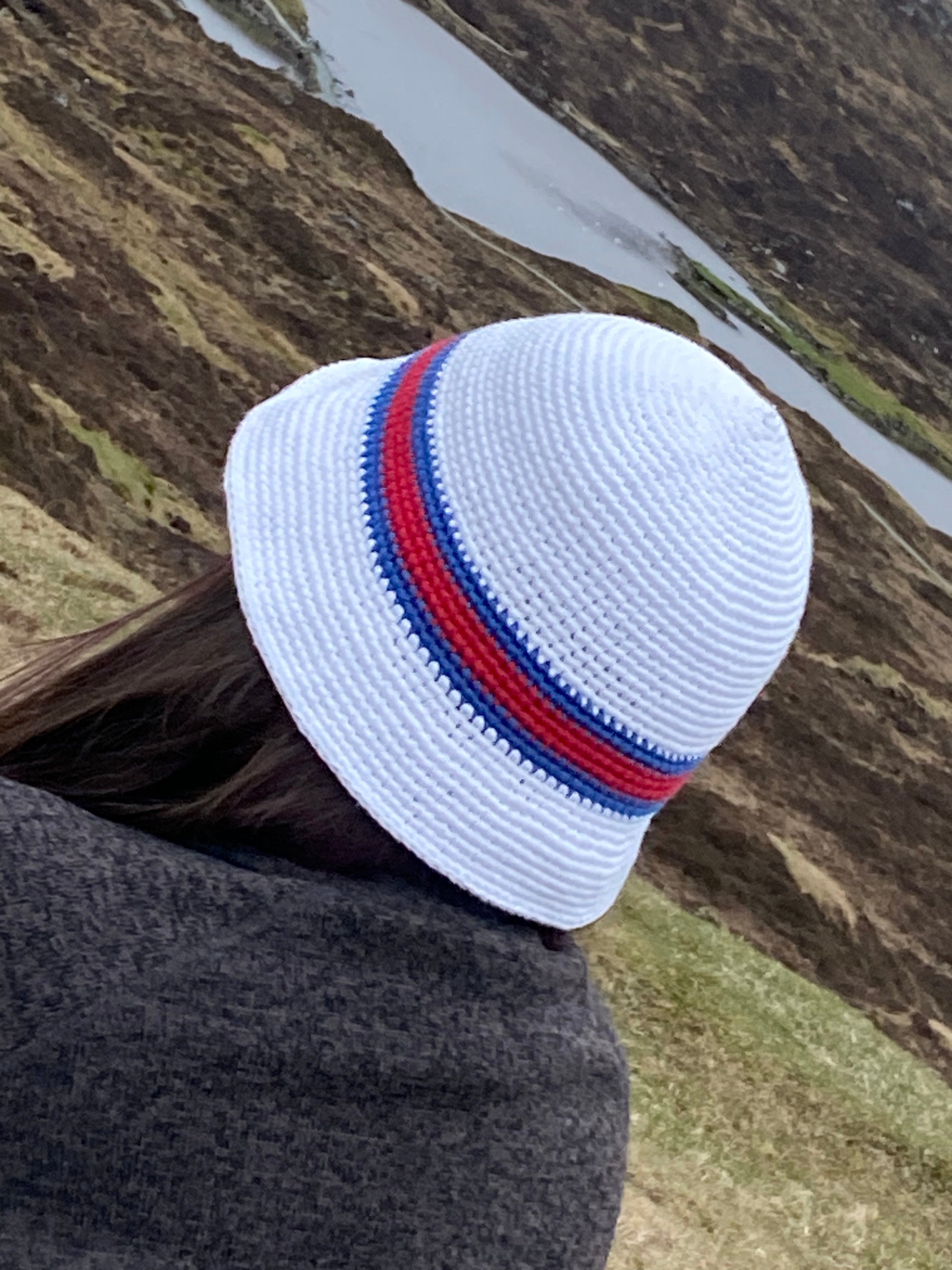 White crocheted hat with red and blue stripes worn by a person, with a scenic background of a river and hills.