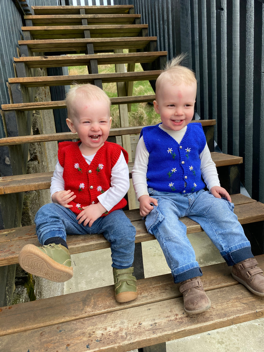 Two young boys sitting on wooden steps, wearing colorful, knitted vests.