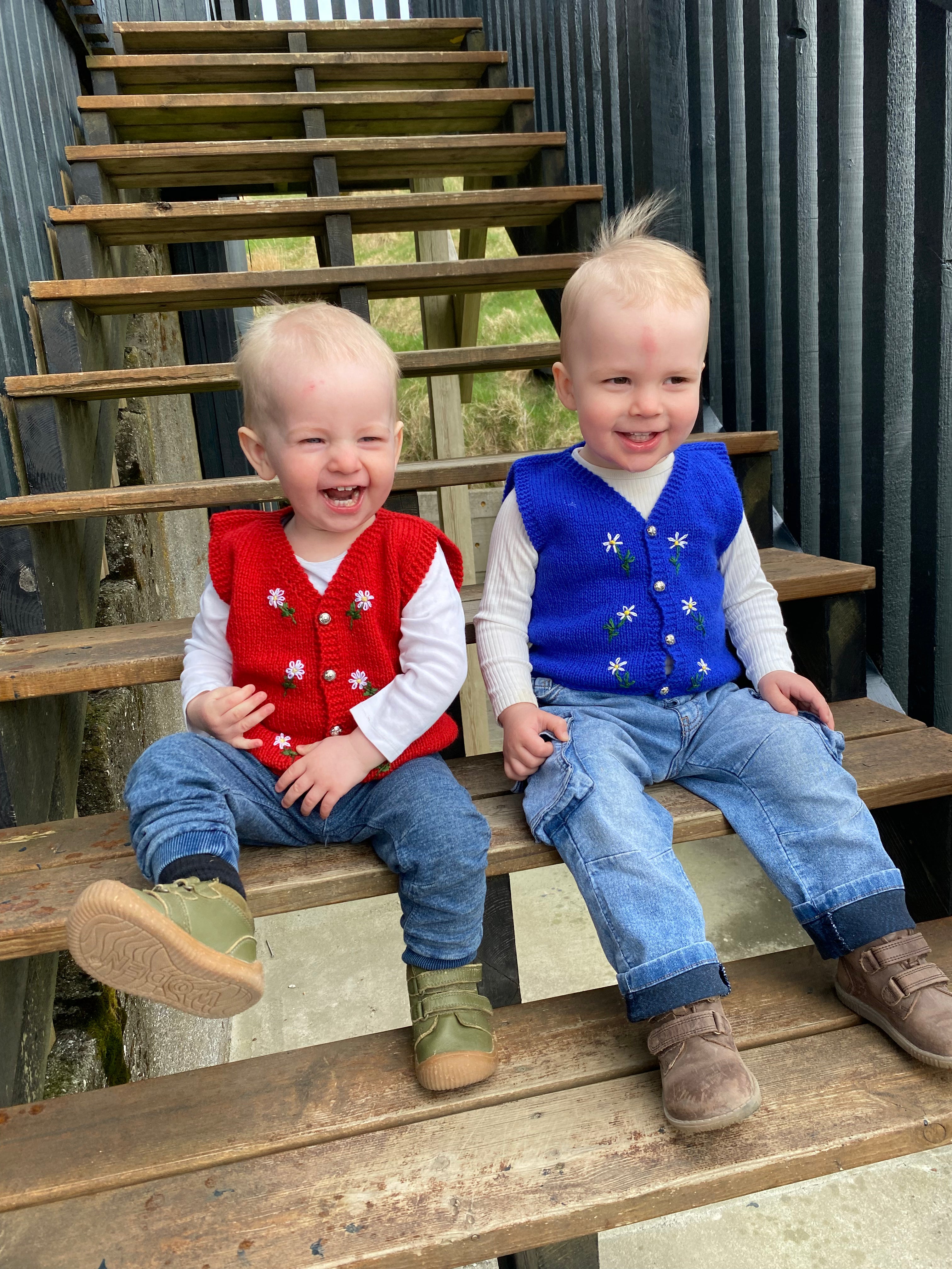 Two young boys sitting on wooden steps, wearing colorful, knitted vests.